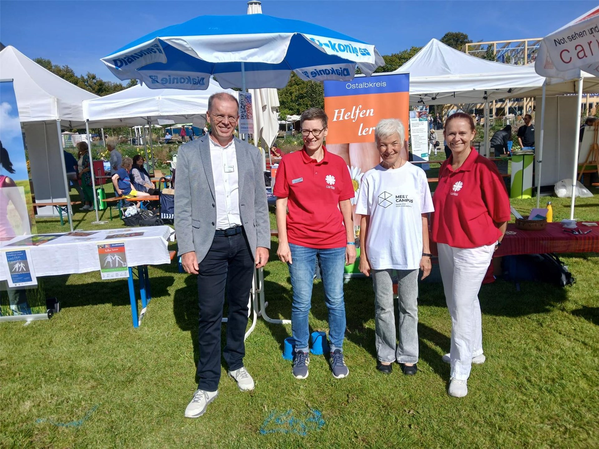 Informationsstand der Caritas beim Ostalbkreis-Aktionstag. Vier Personen stehen vor Pavillons und Infoständen auf einer Wiese. Im Hintergrund sind Besucherinnen und Besucher sowie verschiedene Mitmachangebote zu sehen.
