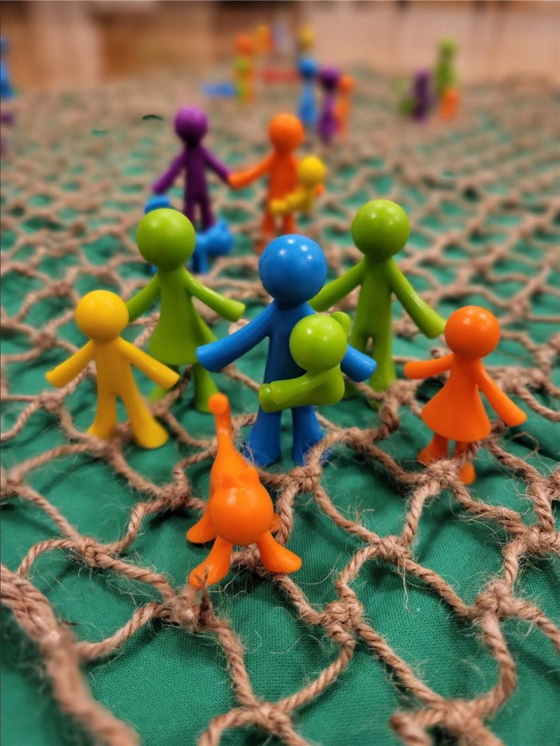 Mehrere bunte Spielfiguren aus Kunststoff stehen auf einem grob geknüpften Seilnetz mit grünem Untergrund. Die Figuren sind in verschiedenen Farben wie Orange, Blau, Grün, Gelb und Lila angeordnet und wirken, als würden sie miteinander interagieren oder sich gegenseitig stützen.